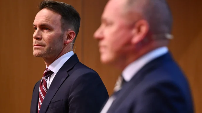 Man in a navy suit and striped tie looking to the left at a formal event; a second man in a suit is blurred in the background.
