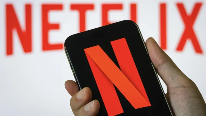 Hand holding a smartphone displaying the red Netflix logo on a black screen, with a blurred Netflix wordmark in the background.