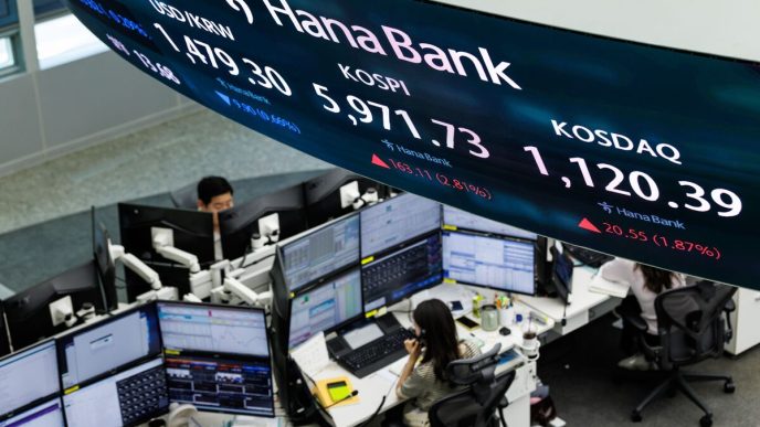 Bank trading floor with multiple computer monitors and traders, beneath a curved Hana Bank stock ticker signage displaying stock prices.