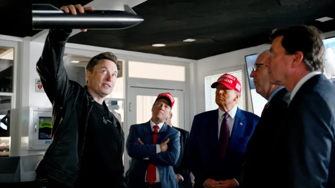 Man in a black jacket raises a rocket-shaped model above his head while four men in suits observe; two wear red MAGA hats.