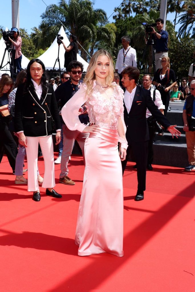 Woman in a pink satin gown posing on a red carpet at a media event with photographers in the background.