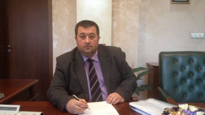 Man in a dark suit and striped tie sits at a wooden desk in an office, signing a document with a pen.
