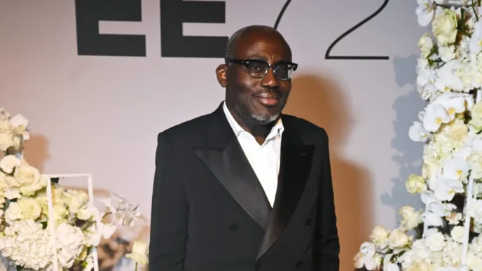 Man in a black tuxedo with satin lapels and a white shirt, wearing glasses, posing at a formal event with white floral arrangements behind him.