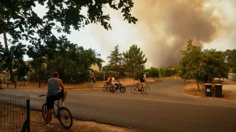 Cyclists gather on a suburban street as a large smoke plume rises from a distant fire in the background.