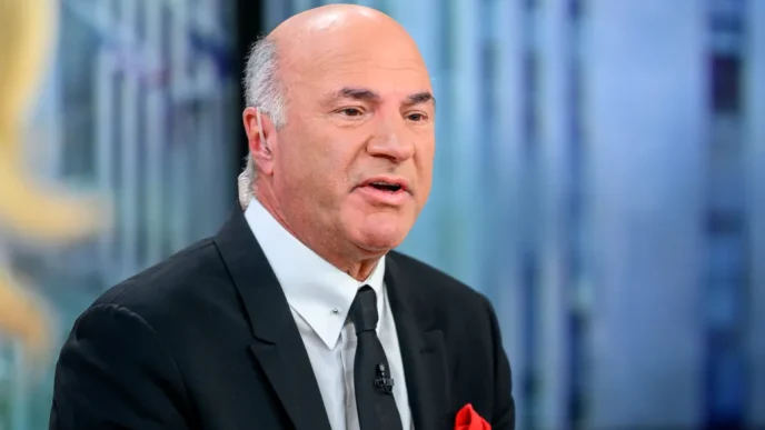 Bald man in a dark suit and white shirt speaking on a TV interview set with a microphone clipped to his lapel and a red pocket square visible.