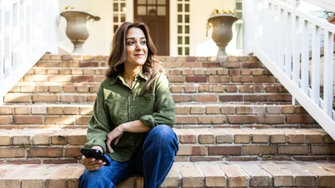 Woman sits on brick stairs outside a house, holding a smartphone and looking to the side, wearing a green shirt and blue jeans.