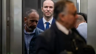 Tall man in a blue suit and tie stands in an elevator, facing the camera, surrounded by blurred passengers around him.