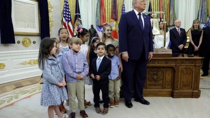 Group of children standing beside a tall man in a dark suit in a ceremonial room with flags and ornate decoried woodwork desk behind them.