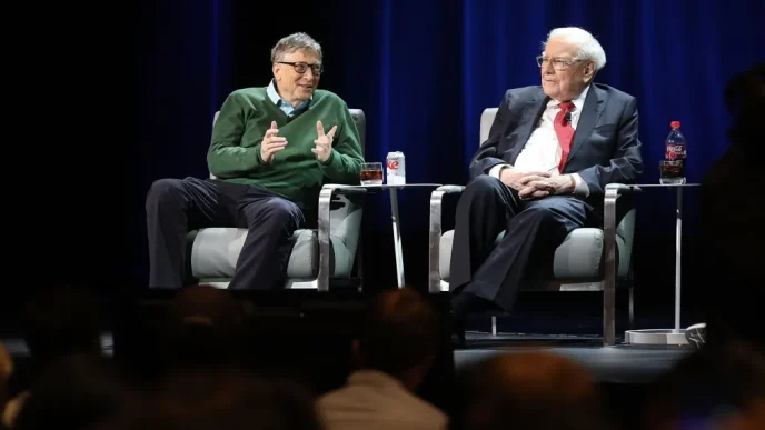 Two men sit in armchairs on stage during a panel discussion; one in a green sweater gestures while the other in a dark suit listens, with drinks on small tables between them and a blue curtain backdrop behind.