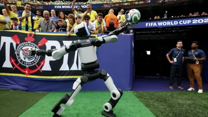 Humanoid robot juggling a soccer ball on a green carpet as cheering stadium crowds watch at a FIFA World Cup event, banner in the background.