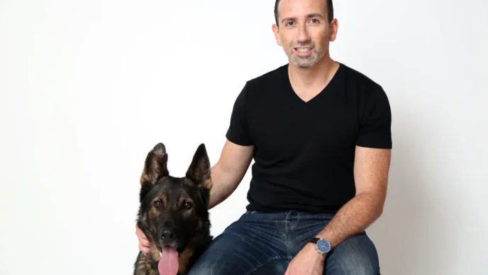 Man in a black V-neck shirt sits next to a German Shepherd with its tongue out, both looking at the camera inside a studio.