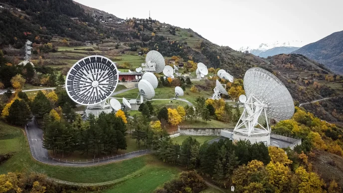 Hilltop complex of large white radio telescopes and dishes amid autumn trees with distant mountains in the background.