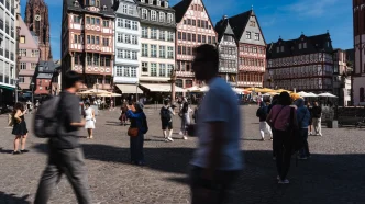 Busy town square with colorful timber-framed buildings lining a cobblestone street, outdoor cafés, and pedestrians in motion under a blue sky.