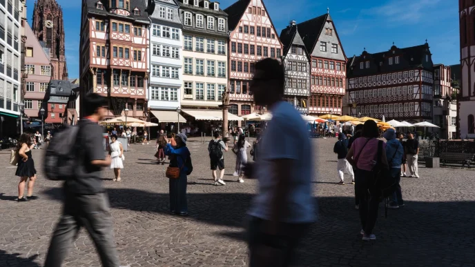 Busy town square with colorful timber-framed buildings lining a cobblestone street, outdoor cafés, and pedestrians in motion under a blue sky.
