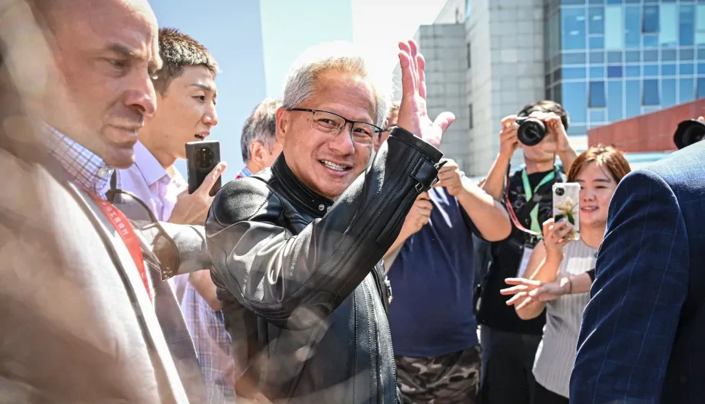 Smiling man with gray hair and glasses waves to a crowd as photographers film him outdoors near a modern building.