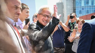 Smiling man with gray hair and glasses waves to a crowd as photographers film him outdoors near a modern building.