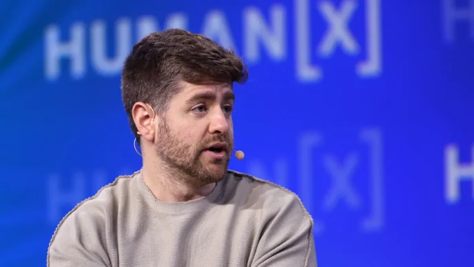 Man with short dark hair and stubble speaks on stage wearing a headset microphone, with a blue backdrop.