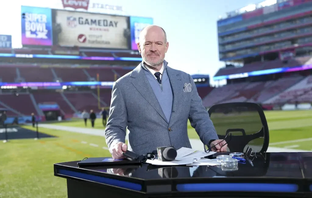 Rich Eisen standing at a broadcast desk inside a football stadium